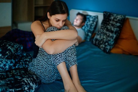 Sad wife sitting on bed edge, holding her legs after fight, her husband is lying in bed in background, his face is lit by cell phone. Sad wife sitting on bed edge, holding her legs after fight, her husband is lying in bed in background, his face is lit by cell phone.