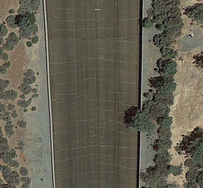 Fig 1. Serious Design Flaw of thinning & weakening of the spillway concrete slabs from Drain pipe within the slabs. This has caused over 5 miles of cracks to form as seen in the 'herringbone' pattern of drains. Image reveals the cracks and repeated patching as whitish strips. This image is over the area of the spillway that 'broke in half'. Image courtesy of Google Earth.